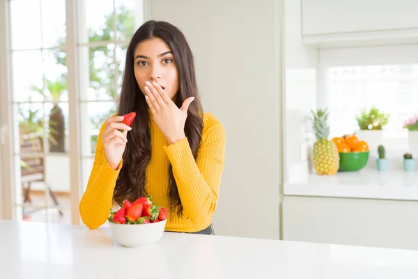 Young woman eating fresh red strawberry as healthy snack cover mouth with hand shocked with shame for mistake, expression of fear, scared in silence, secret concept