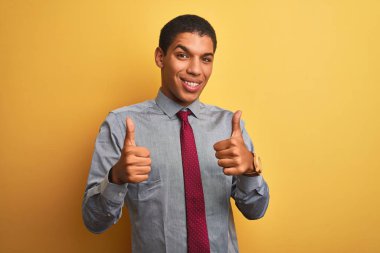 Genç yakışıklı Arap işadamı gömlek giyen ve izole sarı arka plan başarı işareti üzerinde kravat el ile olumlu jest yapıyor, gülümseyerek ve mutlu başparmak kadar. Neşeli ifade ve kazanan jest.