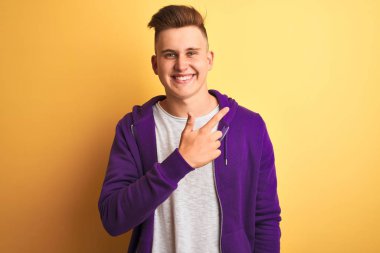 Genç yakışıklı adam mor sweatshirt giyen izole sarı arka plan neşeli bir yüz yüzü ile işaret eden bir gülümseme ile yan yana mutlu ve doğal ifade ile