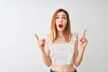Izole beyaz arka plan üzerinde duran rahat t-shirt giyen güzel kızıl saçlı kadın şaşırttı ve yukarı bakarak ve parmakları ile işaret ve kolları kaldırdı şaşırttı.