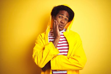 Afro saçlı afro saçlı, izole sarı arka plan üzerinde duran yağmurluk giyen Afrikalı adam yorgun görünümlü ve çapraz kolları ile depresyon sorunları ile sıkılmış düşünme.