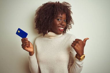 Genç afrikalı afro kadın izole beyaz arka plan üzerinde duran kredi kartı tutan işaret ve mutlu yüzü gülümseyerek yan başparmak ile gösteren