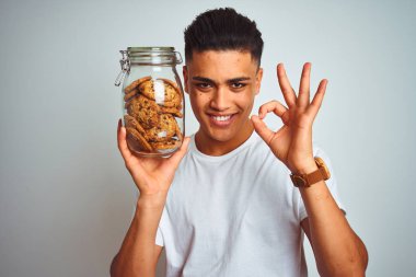 Genç Brezilyalı adam izole beyaz arka plan üzerinde parmakları, mükemmel sembolü ile ok işareti yapıyor ayakta kurabiye kavanozu tutan