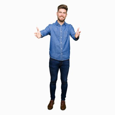 Rahat denim gömlek giyen genç yakışıklı sarışın adam sarılmak için açık kollar ile gülümseyen kamera bakıyor. Mutluluğu kucaklayan neşeli ifade.