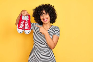 Kıvırcık saçlı genç arap kadın izole sarı arka plan üzerinde rahat spor ayakkabı tutan çok mutlu el ve parmak ile işaret