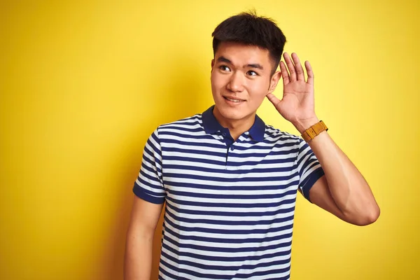 孤立した黄色の背景の上に立つストライプのポロを着た若いアジアの中国人男性は 噂やゴシップに耳を傾けて耳をつんじんで微笑んでいます 聴覚障害の概念 — ストック写真