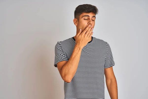 Young indian man wearing navy striped t-shirt standing over isolated white background bored yawning tired covering mouth with hand. Restless and sleepiness.