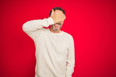 Izole kırmızı arka plan üzerinde gri saçlı yakışıklı orta yaş yaşlı adam gülümseyerek ve sürpriz için gözleri kaplayan yüzünde el ile gülüyor. Kör kavramı.