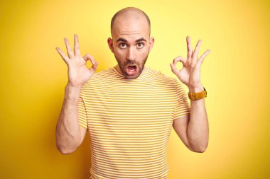 Sakallı genç kel adam sarı izole arka plan üzerinde rahat çizgili t-shirt giyen sürpriz görünümlü ve parmakları ile ok onay sembolü yapıyor şok. Çılgın ifade