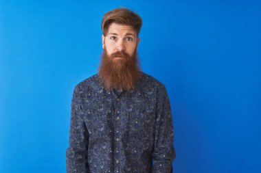 Genç kızıl saçlı İrlandalı adam izole mavi arka plan üzerinde duran çiçek yazlık gömlek giyen yüzünde ciddi ifade ile rahat. Kameraya basit ve doğal bir bakış.