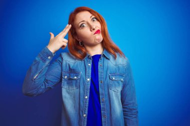 Genç güzel kızıl saçlı kadın mavi izole arka plan üzerinde duran kot gömlek giyen Çekim ve kendini silah gibi baş el ve parmakları gösteren öldürme, intihar jesti.