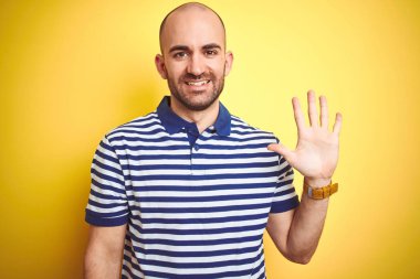 Sakallı genç kel adam sarı izole arka plan üzerinde rahat çizgili mavi t-shirt giyen gösteren ve emin ve mutlu gülümserken parmakları sayısı beş işaret.