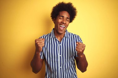 Afro saçlı afro saçlı bir adam izole sarı arka plan üzerinde duran çizgili gömlek giyen çok mutlu ve heyecanlı kolları kaldırdı ile kazanan jest yapıyor, gülümseyerek ve başarı için çığlık. Kutlama konsepti.