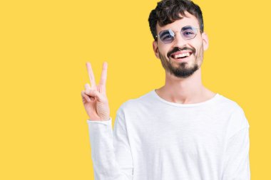 Genç yakışıklı adam mutlu yüz zafer işareti yaparken kameraya göz kırpıyor ile gülümseyen izole arka plan üzerinde güneş gözlüğü takıyor. İki numaralı.
