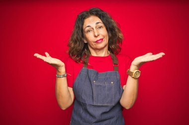 Orta yaş yaşlı kadın kırmızı izole arka plan üzerinde önlük üniforma giyen habersiz ve kollar ve eller kaldırdı ile karışık ifade. Şüphe kavramı.