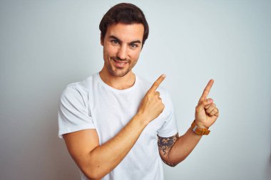 Izole beyaz arka plan üzerinde rahat t-shirt giyen dövme ile genç yakışıklı adam gülümseyerek ve yan iki el ve parmakları ile işaret kamera bakıyor.