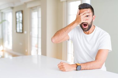 Yakışıklı İspanyol adam evde rahat beyaz t-shirt şok yüz ve gözleri el ile kapsayan gözetleme, utanmış ifade ile parmaklarından bakarak.