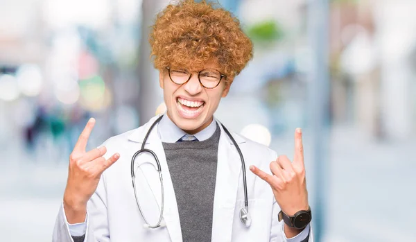 Young handsome doctor man wearing medical coat shouting with crazy expression doing rock symbol with hands up. Music star. Heavy concept.