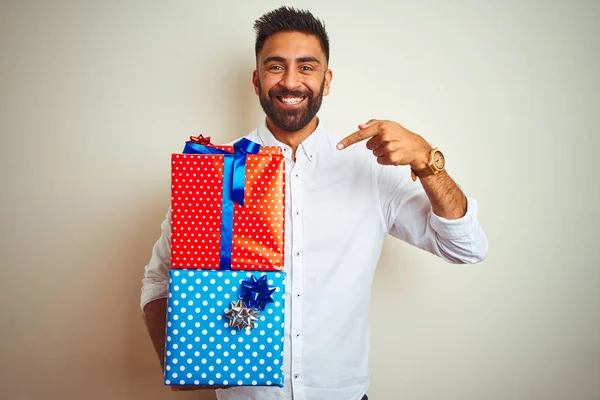 Young arab indian hispanic man holding birthday gifts standing over isolated white background very happy pointing with hand and finger