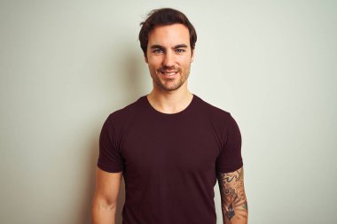 Yüzünde mutlu ve serin bir gülümseme ile izole beyaz arka plan üzerinde mor rahat t-shirt giyen dövme ile genç yakışıklı adam. Şanslı kişi.