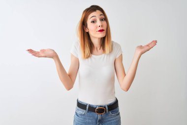 Güzel kızıl saçlı kadın rahat t-shirt izole beyaz arka plan üzerinde duran giyen habersiz ve kollar ve eller kaldırdı ile karışık ifade. Şüphe kavramı.