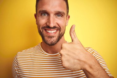 Yakın sarı izole arka plan üzerinde rahat t-shirt giyen genç yakışıklı adam el ile jest kadar mutlu başparmak yapıyor. Başarı göstererek kameraya bakan ifadeyi onaylama.
