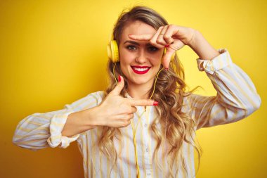 Genç güzel kadın sarı izole arka plan üzerinde kulaklık kullanarak müzik dinlerken mutlu yüz ile elleri ve parmakları ile çerçeve yapma gülümseyerek. Yaratıcılık ve fotoğrafçılık konsepti.