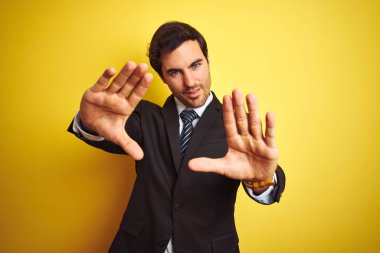 Takım elbise giyen ve kravat giyen genç yakışıklı işadamı izole sarı arka plan üzerinde duran avuç içi ve parmaklar kullanarak çerçeve yapıyor, kamera perspektifi