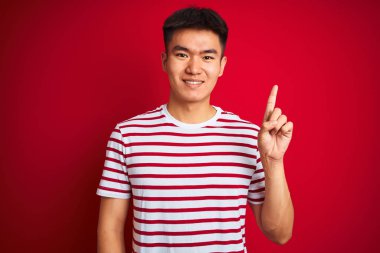 Genç asyalı Çinli adam çizgili t-shirt izole kırmızı arka plan üzerinde duran gösteren ve kendinden emin ve mutlu gülümserken parmak sayısı bir işaret giyen.
