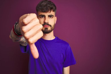 Dövme giyen genç adam izole mor arka plan üzerinde duran t-shirt mutsuz ve kızgın gösteren reddi gösteren ve aşağı jest başparmak ile negatif. Kötü ifade.