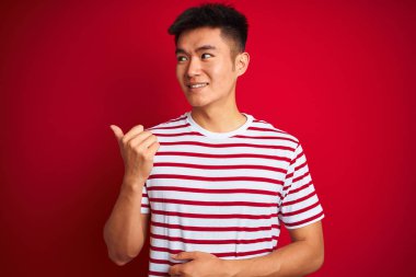 İzole kırmızı arka plan üzerinde duran çizgili t-shirt giyen genç Asyalı adam mutlu yüz görünümlü ve başparmak ile yan işaret gülümseyerek.