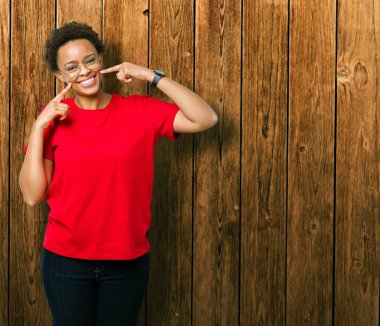 Kendinden emin gösterilen ve işaret parmakları diş ve ağız ile gülümseyen izole arka plan üzerinde gözlük güzel genç Afro-Amerikan kadın. Sağlık kavramı.