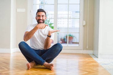 Yakışıklı İspanyol adam evde katta oturan rahat t-shirt giyen hayran ve el ile sunarken kameraya gülümseyerek ve parmak ile işaret.