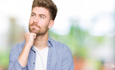 Genç yakışıklı sarışın adam el çenesi ile rahat gömlek giyiyor soru hakkında düşünme, dalgın ifade. Düşünceli bir yüz ile gülümsüyor. Şüphe kavramı.