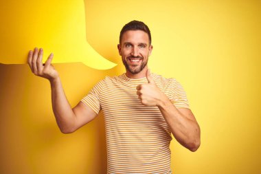 Genç adam ok işareti yapıyor büyük bir gülümseme ile mutlu sarı izole arka plan üzerinde mesaj için boş konuşma balonu tutan, parmaklarla başparmak, mükemmel işareti