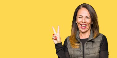 Mutlu yüz zafer işareti yaparken kameraya göz kırpıyor ile gülümseyen kış yelek giyen güzel orta yaş kadın. İki numaralı.