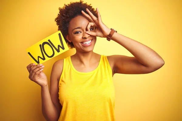 Young african american woman showing wow banner board over yellow isolated background with happy face smiling doing ok sign with hand on eye looking through fingers