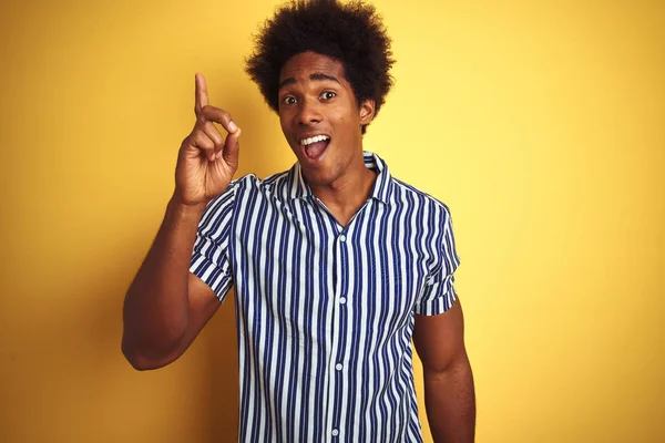 American man with afro hair wearing striped shirt standing over isolated yellow background pointing finger up with successful idea. Exited and happy. Number one.