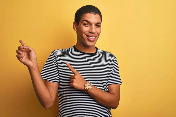 年轻的英俊阿拉伯男子穿着海军条纹T恤在孤立的黄色背景微笑 看着相机指着两只手和手指的一侧 — 图库照片
