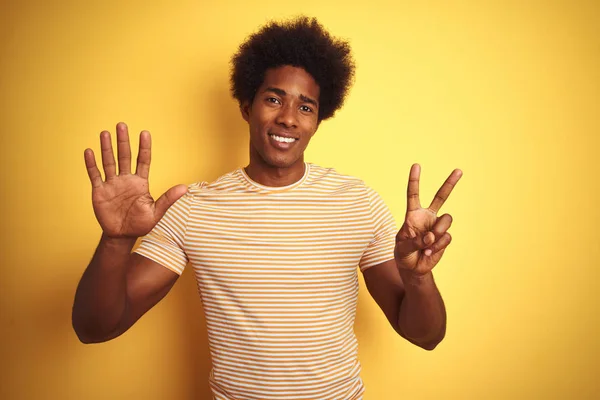 孤立した黄色の背景の上に立つストライプのTシャツを着たアメリカ人男性は 自信を持って幸せに微笑みながら 指番号7を示し 指を上げている — ストック写真