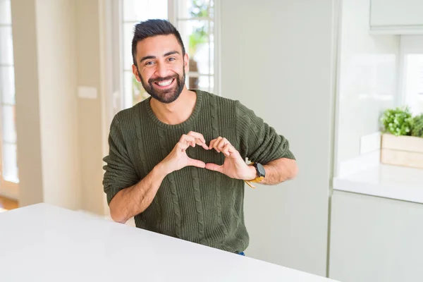 カジュアルなセーターを着て家でハンサムなヒスパニック男恋ハートマークと手で形を示す笑みを浮かべてください ロマンチックな概念 — ストック写真