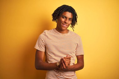 Dreadlocks ile afro adam birlikte elleri ile izole sarı arka plan üzerinde duran çizgili t-shirt giyen ve rahat ve neşeli gülümseyen parmakları geçti. Başarı ve iyimser