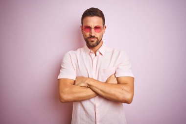 Yakışıklı adam pembe izole arka plan şüpheci ve sinir üzerinde zarif yazlık gömlek ve güneş gözlüğü giyen, çapraz kolları ile yüzünde onaylamayan ifade. Negatif kişi.