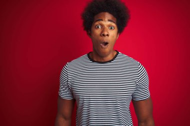 Afro saçlı genç amerikalı adam izole kırmızı arka plan üzerinde lacivert çizgili t-shirt giyen korkmuş ve sürpriz ifade, korku ve heyecanlı yüz ile şok.