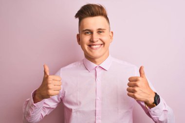Genç yakışıklı işadamı izole pembe arka plan başarı işareti el ile olumlu jest yapıyor ayakta zarif gömlek giyen, gülümseyerek ve mutlu başparmak kadar. Neşeli ifade ve kazanan jest.