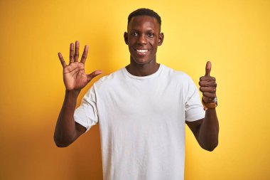 Beyaz tişört giyen genç afro-amerikan adam izole sarı arka plan üzerinde duran gösteren ve parmakları ile işaret ederken kendinden emin ve mutlu gülümseyerek altı numara.