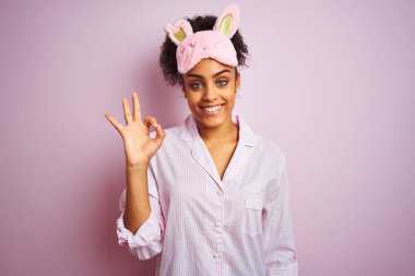 Genç afro-amerikan kadın izole pembe arka plan üzerinde pijama ve maske giyen olumlu el ve parmakları ile ok işareti yapıyor gülümseyerek. Başarılı ifade.
