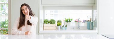 Güzel genç kadının evde beyaz masada oturan geniş açı resim el ile jest kadar mutlu başparmak yapıyor. Başarı göstererek kameraya bakarak ifadeyi onaylama.