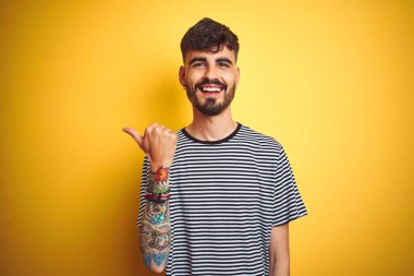Çizgili t-shirt giyen dövme giyen genç adam izole sarı arka plan üzerinde mutlu yüz görünümlü gülümseyerek ve başparmak ile yan işaret.