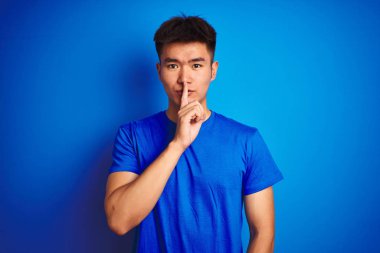 Young asian chinese man wearing t-shirt standing over isolated blue background asking to be quiet with finger on lips. Silence and secret concept. clipart